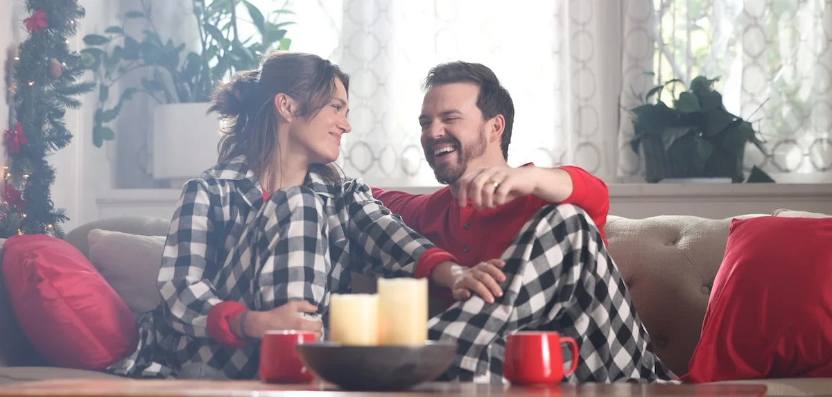 {What's Included in|What Happens During|What’s} a Heating Tune-Up (and When {Should You|is the Best Time to} Schedule One?) 2 Couple sitting on a couch laughing in matching buffalo plaid pajamas with red mugs and Christmas greenery hung in the background